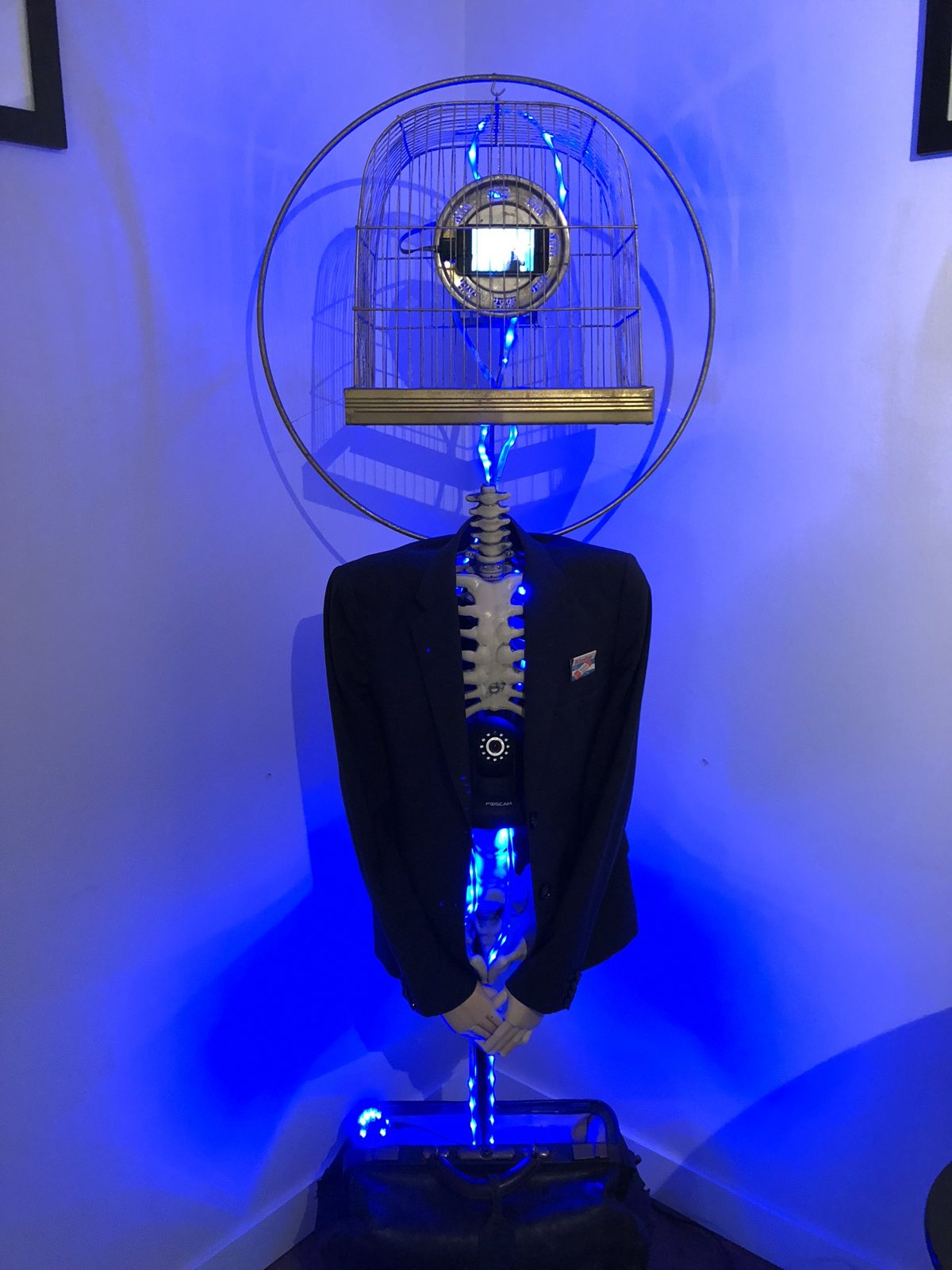 A birdcage with a small screen inside sits above a suit jacket and a lit spine, ringed by a hoop, on an old doctor’s bag, all washed in blue light