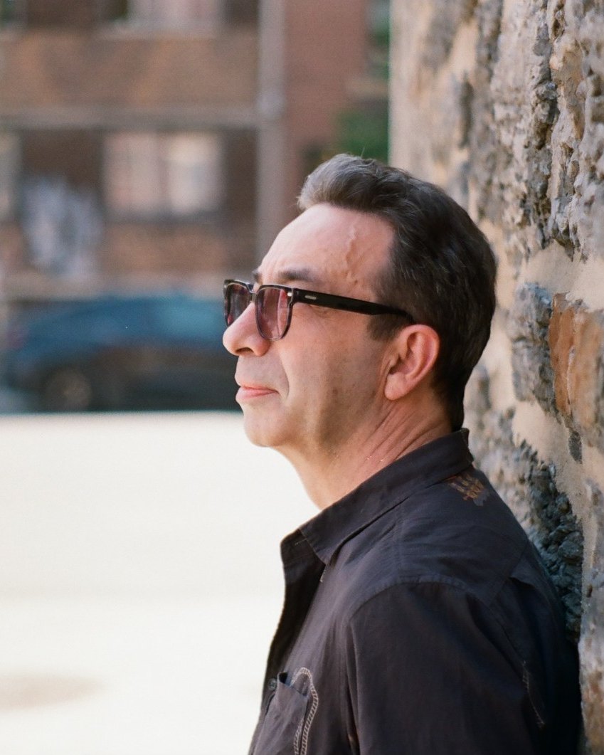 Dion Le Roux in profile, wearing dark glasses, leaning against a stone wall on a city street